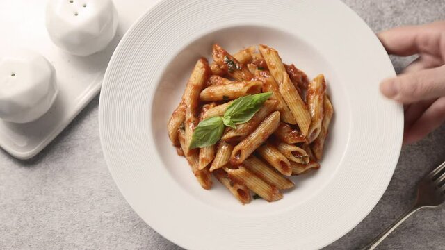 The girl sets and clears a plate of penne pasta, tomato sauce and basil from the table. Light background, close-up 