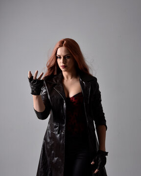 Close up portrait of girl with long red hair wearing dark leather coat, corset and gloves. Posing with gestural hand movements as if casting spell. Moody back lit lighting against a studio background.
