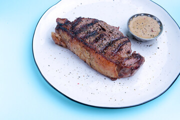 Delicious beef steak with sauce served on a white plate over pastel blue background. BBQ meat, barbecue concept.