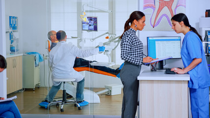 Obraz premium Patients asking informations filling in dental document preparing for teeth exemination. Senior woman sitting on chair in waiting area of crowded orthodontist office while doctor working in background