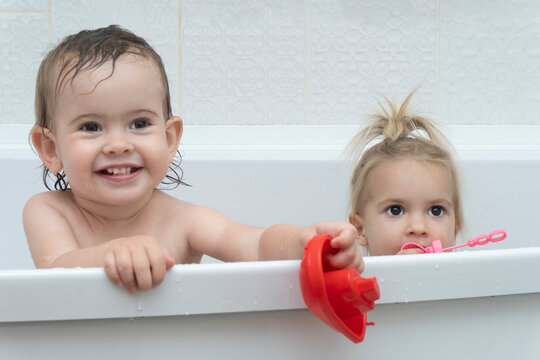 Portrait of two little caucasian cute girl sisters sitting at bathtub, having time at taking bath, playing with bath toys, smiling, white background, copy space. 