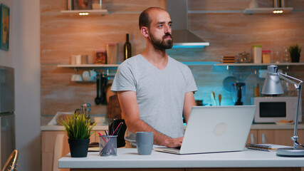 Man thinking at next project while working from home reading the tasks looking on laptop sitting in home kitchen. Busy employee using modern technology network wireless doing overtime reading writing