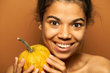 Pumpkin mania. Close up portrait of excited young mixed race woman smiling at camera, holding little pumpkin, posing isolated over orange background. Healthy eating, skincare concept