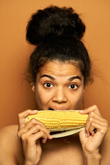 Bite it. Portrait of adorable young mixed race woman looking at camera, holding and biting yellow corn cob, posing over orange background. Healthy eating, vegetarian concept