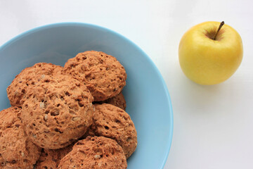 Homemade oatmeal cookies in a blue bowl and golden apple on white table. Overhead view of oat biscuits and fresh fruits on white background with copy space. 