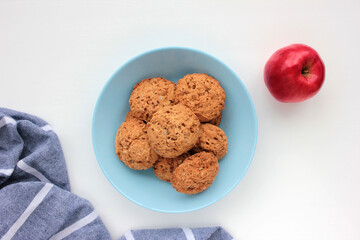 Homemade oatmeal cookies in a blue bowl and red apple on white table.Flat lay composition with oat biscuits, fresh fruit, and gray napkin on white background with copy space.