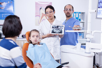 Pediatric dentist discussing about cavity prevention with parent of little girl showing x-ray on tablet. Stomatologist explaining teeth diagnosis to mother of child in health clinic holding x-ray.