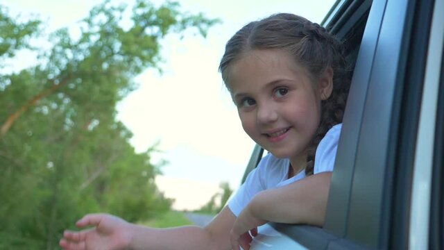 A happy child travels with his parents by car. A cheerful sweet girl climbed out of the car window, smiles and waves. The concept of a happy family, the path to a dream, freedom.