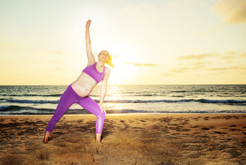 Pregnant woman exercise stretching on the sand beach with sunset over ocean on background