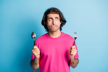 Portrait of attractive minded guy holding in hands fork spoon licking lip counting calories...