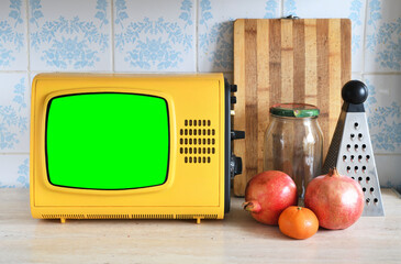 An old yellow vintage TV with a green screen for adding videos and images stands in the kitchen next to fruits and kitchen utensils against the backdrop of ceramic tiles.