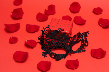 Valentine's day black lace mask andgiftbox with gold hearts on red background with rose petals