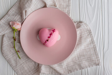 Fototapeta na wymiar Pink heart-shaped mousse cake on a pink round plate next to a rosebud on a white wooden background and pastel fabric. Congratulations on Valentine's Day, Women's Day