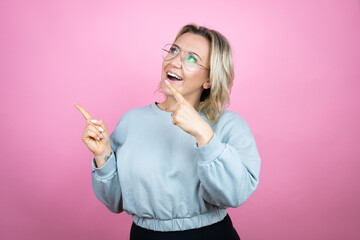 Young caucasian woman wearing sweatshirt over pink background surprised and pointing her fingers side