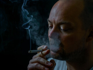 Fototapeta premium Man smoking a cigarette blowing out white smoke. Isolated on black background, shallow depth of field.