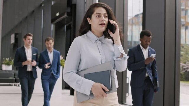 Medium shot of young mixed-race businesswoman wearing blue shirt going along office hall and having telephone call to colleague