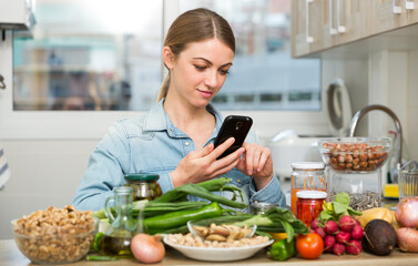 Portrait of glad woman looking at mobile phone while making dish on kitchen at home. High quality photo