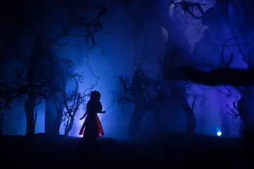 Horror Halloween decorated conceptual image. Alone girl with the light in the forest at night. Silhouette of girl standing between trees with surreal light on background. Selective focus.