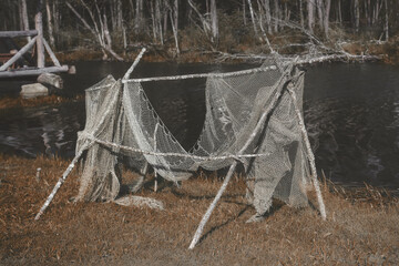 Antique fishnet for fishing hanging on framework made from wooden sticks among dry weathered grass on the shore of a lake. Outdated fishing gear on natural background. Toned