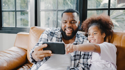 Happy African American family dad and daughter having fun and using mobile phone video call on sofa at house. Self-isolation, stay at home, social distancing, quarantine for coronavirus prevention.