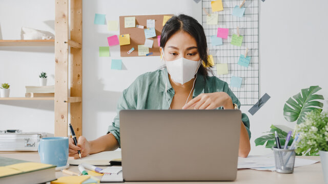 Asia businesswoman wearing medical face mask using laptop talk to colleagues about plan in video call while working from home at living room. Social distancing, quarantine for corona virus prevention.
