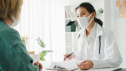 Young Asia lady doctor wear protective mask using clipboard is delivering great news talk discuss results or symptoms with girl patient in hospital office. Lifestyle new normal after corona virus.