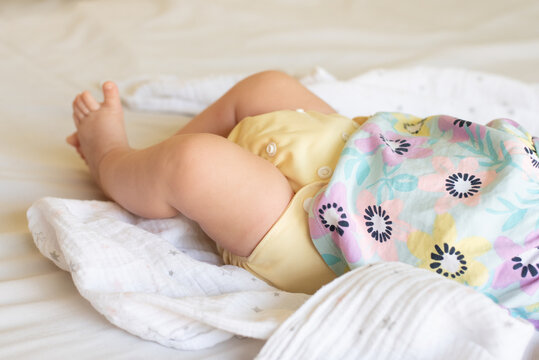 A baby girl lying on a bed on linen wearing a yellow cloth, reusable diaper and a pretty dress with flowers.