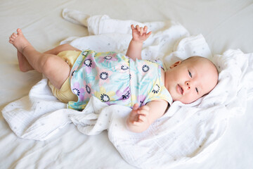 A baby girl lying on a bed on linen wearing a yellow cloth, reusable diaper and a pretty dress with flowers.