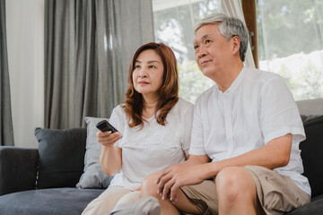 Obraz premium Asian elderly couple watching television in living room at home, sweet couple enjoy love moment while lying on the sofa when relaxed at home. Enjoying time lifestyle senior family at home concept.
