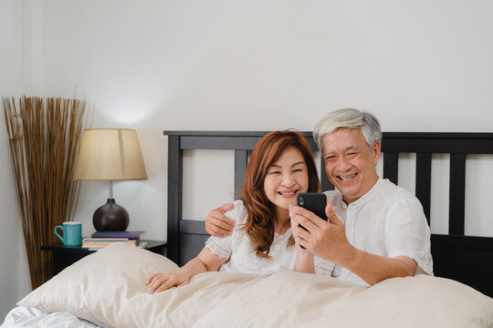 Asian senior couple selfie at home. Asian Senior Chinese grandparents, husband and wife happy using mobile phone selfie after wake up lying on bed in bedroom at home in the morning concept.