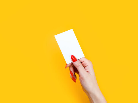 Female Hand With Red Manicure Gives Blank White Business Card, Close-up, Isolated On Yellow Background. Mockup