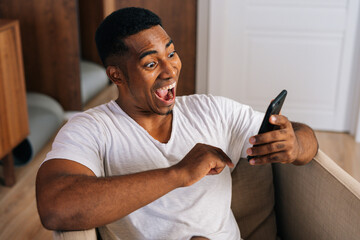 Close-up face of surprised African-American man receiving good news using mobile phone at home. Shocked male with open mouth enjoying smartphone communication looking with amazement at message on cell