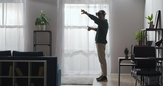 man is viewing photos and pictures by hmd gadget in apartment, zooming by hands movement, standing alone in living room