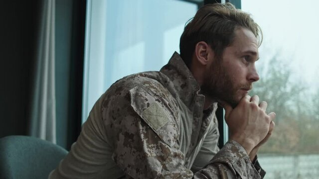 A concentrated young american soldier man is looking to the window sitting inside the apartments