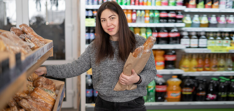 A brunette stands at the shelves in the store, smiling and holding wheat bread in a paper bag. Woman's look.
