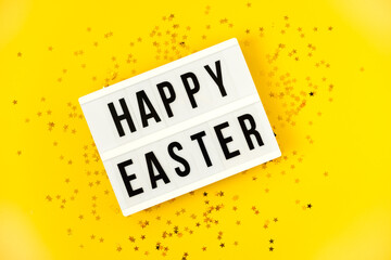 Light box over bright yellow background with the text Happy Easter. Easter celebration concept. Golden eggs and stars confetty