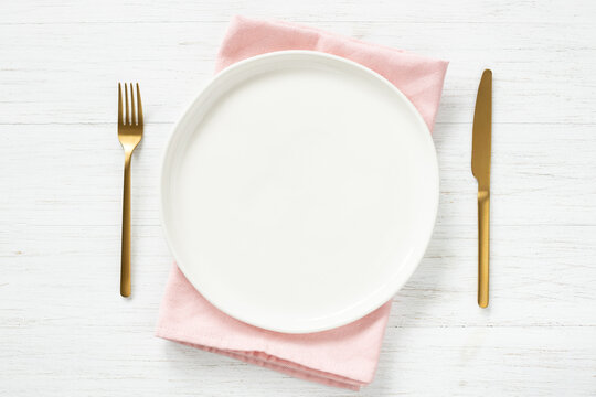 White empty plate on pink napkin and golden cutlery on wooden table. Top view on table setting.