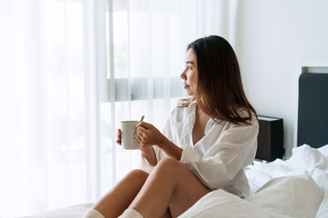 Young beautiful brunette hair girl in white shirt pajamas drinking coffee while sitting on the bed in the morning.