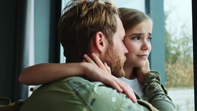 A close-up view of a calm soldier man is hugging with his crying young daughter sitting at home and looking to the window