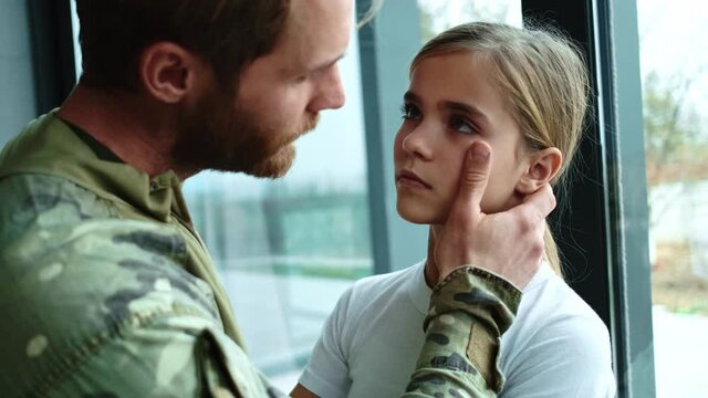 A close-up view of a sad soldier man is looking at his young daughter sitting at home before leaving