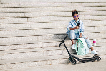 Stylish beautiful young woman sitting on steps after riding on electric scooter and texting friends or checking social media © DragonImages