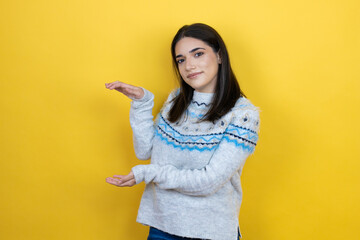 Young caucasian woman wearing casual sweater over yellow background gesturing with hands showing big and large size sign, measure symbol. Smiling . Measuring concept.