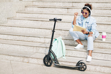 Happy stylish young woman in sunglasses sitting on steps and watching show on digital tablet after riding on electric scooter on campus © DragonImages
