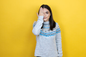 Young caucasian woman wearing casual sweater over yellow background covering one eye with hand,...