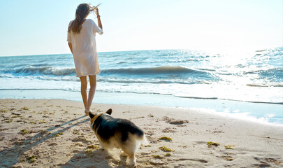 Back view woman in oversize shirt standing by sea at coastline with morning sunlight, cute dog next to owner enjoying seaview together