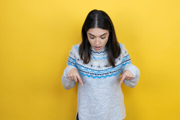 Young caucasian woman wearing casual sweater over yellow background surprised, looking down and pointing down with fingers and raised arms