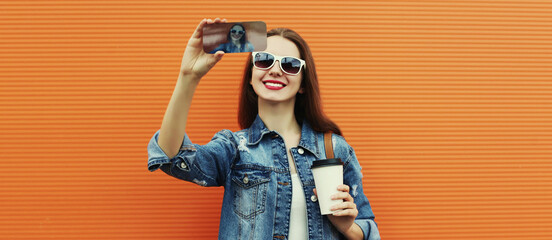 Portrait of smiling woman taking a selfie picture by smartphone wearing a denim jacket on a orange background