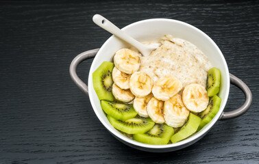 Oatmeal porridge with bananas, kiwi in a bowl with a spoon on a dark background with copy space close up. Healthy breakfast