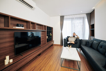 Handsome software developer sitting on armchair in his apartment, working on laptop and thinking over next line of code