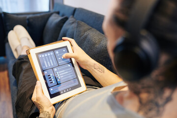 Man in headphones lying on sofa and controlling volume of music via application on tablet computer